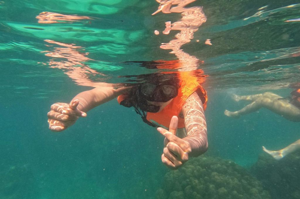 오렌지색 구명조끼를 입고 양손으로 엄지손가락을 치켜세운 표지판을 보여주며 수중에서 스노클링을 하는 사람.