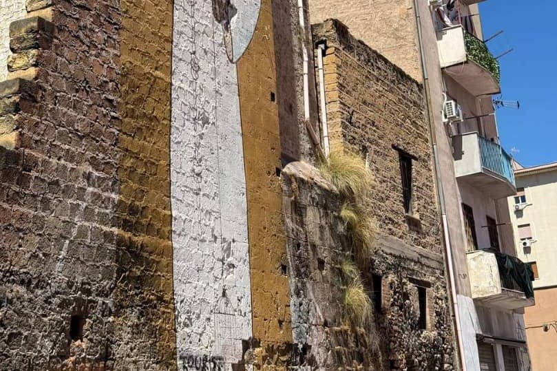 벽에 부분 벽화가 있고 틈새에서 자라는 잔디가 있는 오래된 석조 건물. 눈에 띄는 옥상과 바가 있는 창문.