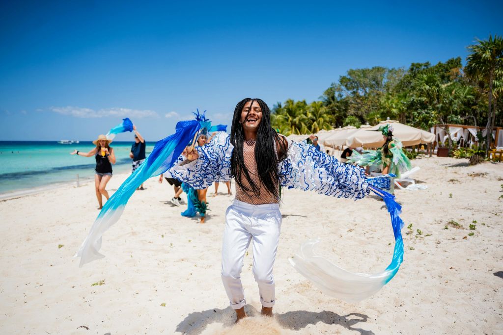 한 사람이 맑고 푸른 하늘 아래 모래 해변에서 파란색과 흰색 천 스트립을 들고 다른 사람들과 함께 즐겁게 춤을 추고 있습니다.