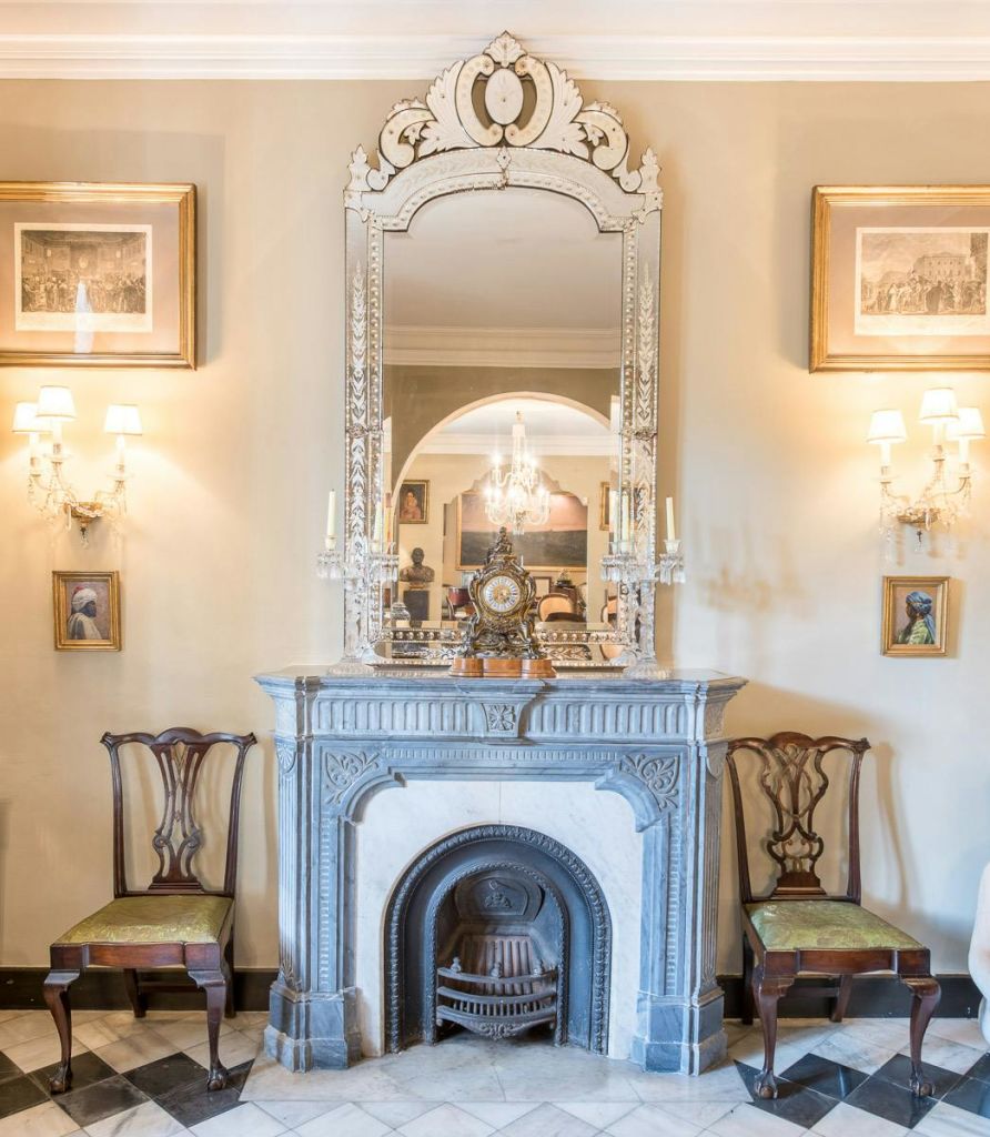 화려한 벽난로, 커다란 거울, 의자 2개, 벽 스콘, 액자에 담긴 예술품이 있는 우아한 객실로, 샹들리에가 반사되어 있습니다.