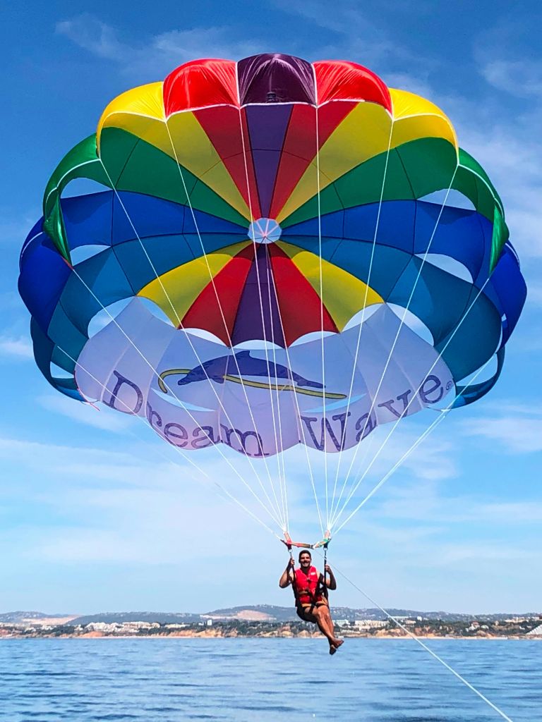 맑고 푸른 하늘과 먼 풍경을 배경으로 "꿈의 물결" 이라는 라벨이 붙은 커다란 다색 낙하산을 타고 패러세일링을 하는 사람.