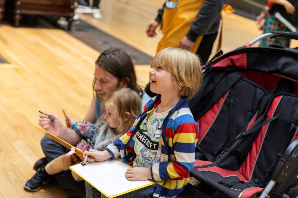 공책을 든 어린이 두 명과 어른 한 명이 나무 바닥에 앉아 있습니다. 한 아이는 연필을 들고, 다른 아이는 미소를 짓고, 어른은 아래를 내려다봅니다.