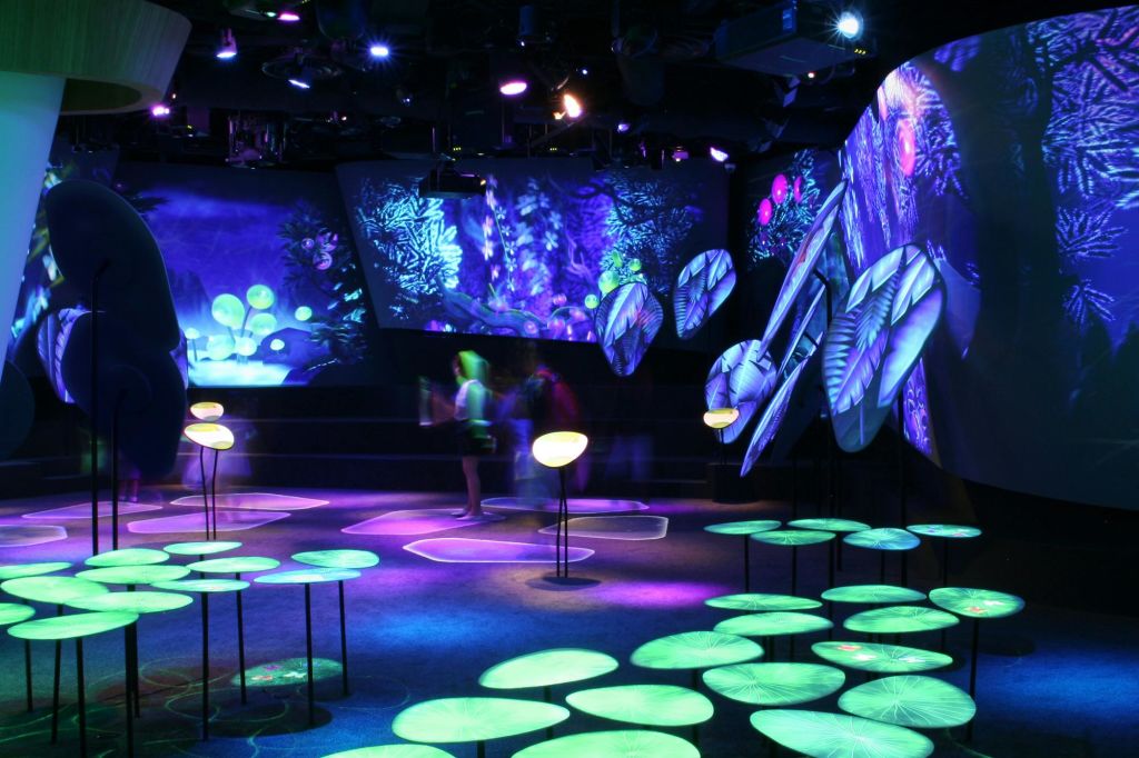 원형 조명 설치물과 추상적인 디자인이 특징인 식물과 곰팡이의 네온 조명 투영이 있는 어두운 방. 배경에 흐릿한 형상이 있습니다.