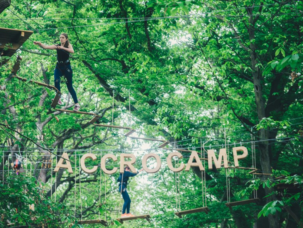 울창한 숲에서 나무 꼭대기 로프 코스를 항해하는 두 사람, "ACCROCAM" 이라는 큰 글자가 전면에 매달려 있습니다.