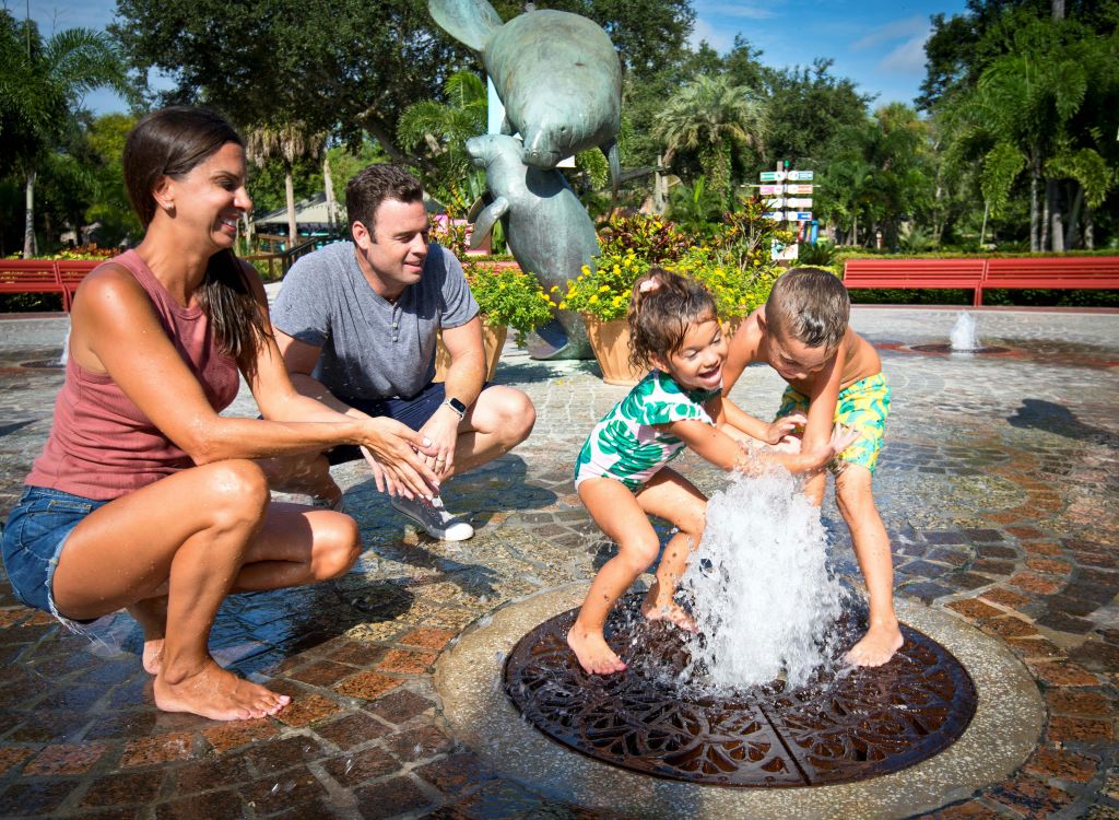 성인 2명과 어린이 2명이 분수대에서 물 뿌리기 체험을 하고, 조각상과 녹음이 우거진 분수를 배경으로 물놀이를 즐깁니다.