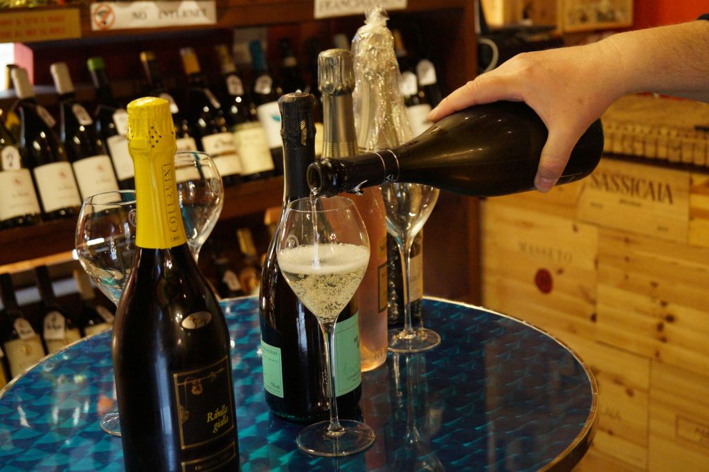 샴페인 병과 와인 랙 배경으로 둘러싸인 파란색 테이블 위에 놓인 유리잔에 손으로 스파클링 와인을 붓습니다.