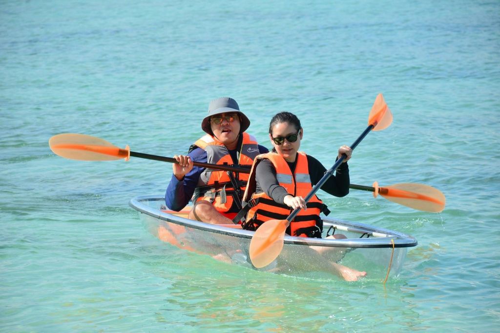 구명조끼를 입은 두 사람이 푸르고 잔잔한 바다에서 투명 카약을 타고 있습니다. 둘 다 선글라스를 착용하고 있으며, 한 명은 모자를 쓰고 있습니다.