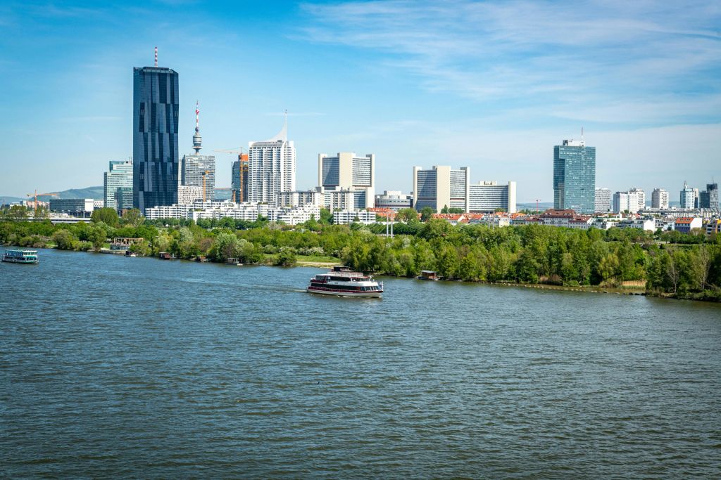 현대적인 고층 건물, 강 위의 보트, 맑고 푸른 하늘 아래 강둑을 가로지르는 나무가 있는 도시 스카이라인.