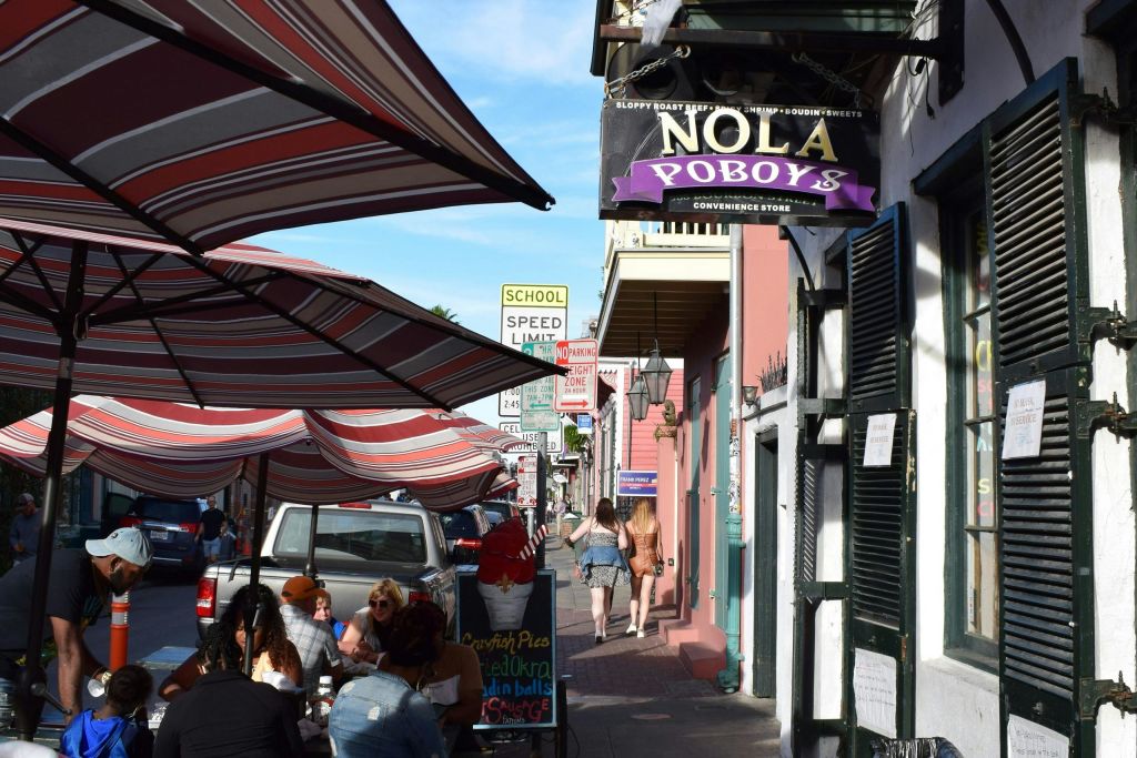 '놀라 포보이 (NOLA Poboys)' 라고 적힌 간판이 있는 상점 근처의 줄무늬 우산 아래에서 야외 식사를 즐기는 사람들이 있는 거리 풍경.
