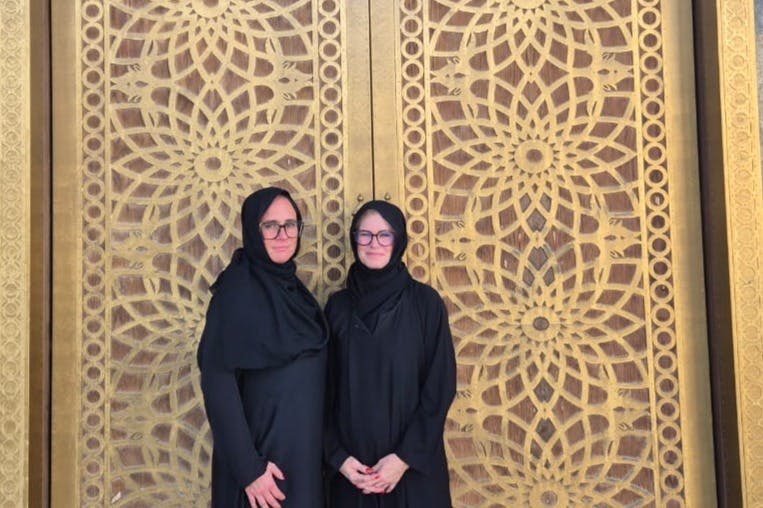 검은 복장과 머리 스카프를 두른 두 사람이 정교한 기하학적 무늬가 있는 화려하고 황금빛 문 앞에 서 있습니다.