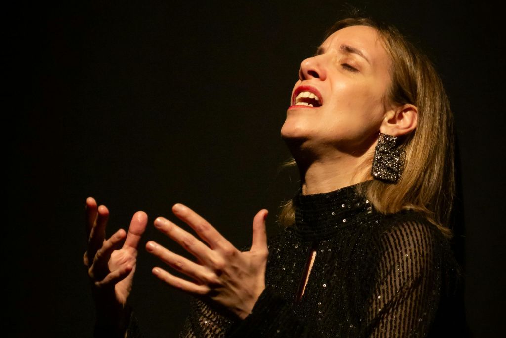 눈을 감고 입을 벌린 채 스팽글과 커다란 장식 귀걸이가 달린 검은색 의상을 입고 어두운 배경에 대고 있는 사람.