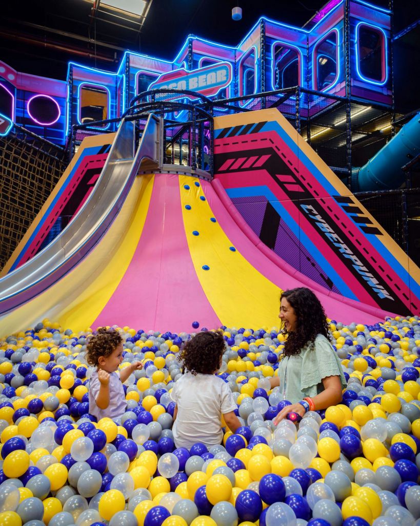 실내 놀이터에서 화려한 미끄럼틀 앞에서 노란색, 파란색, 흰색 공을 들고 볼 구덩이에서 놀고 있는 세 사람.