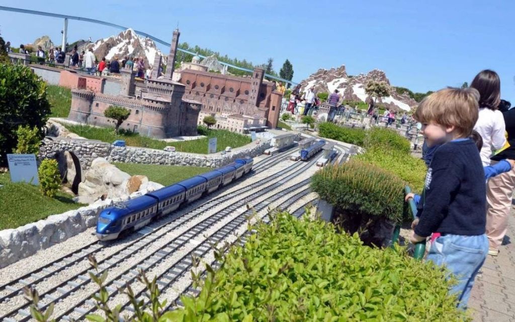 녹지로 둘러싸인 도시의 상세한 스케일 모델로 트랙을 달리는 미니어처 열차. 사람들이 디스플레이를 관찰하고 있습니다.