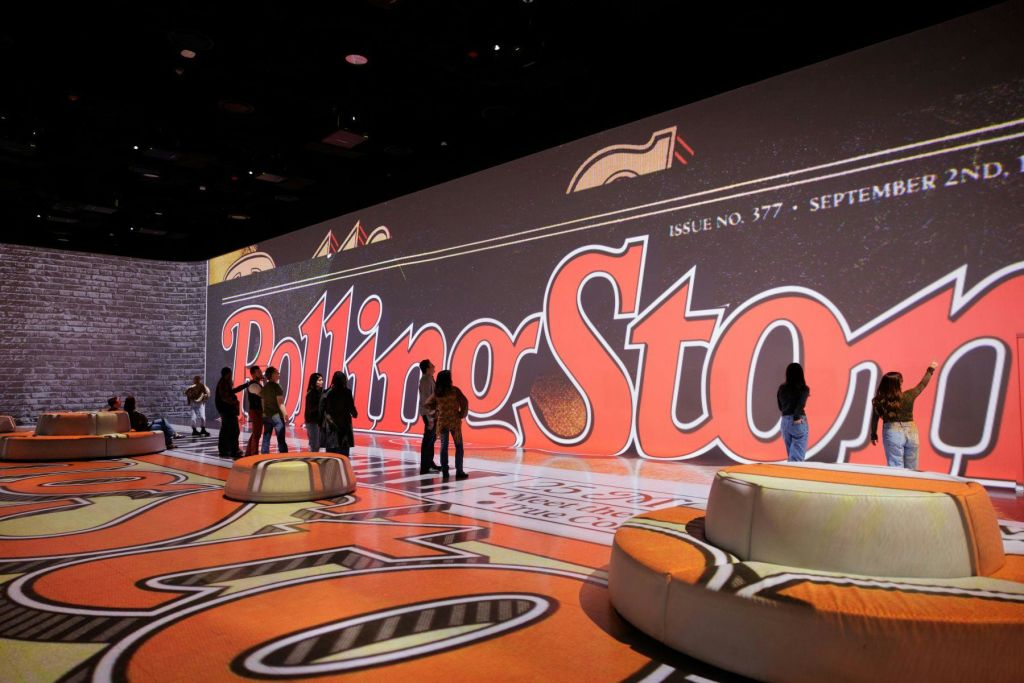 대형 벽과 바닥에 "롤링 스톤" 이 인쇄된 큰 방에 사람들이 서서 걷고 있으며, 일부 좌석 공간이 보입니다.