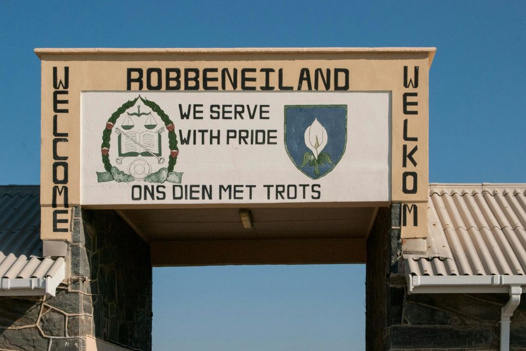 Robben Island의 입구 표지판 옆에는 "We SERVE WITH PRIDE/ONS DIEN MET TROTS" 및 "WELKOM" 이라는 문구가 있습니다.