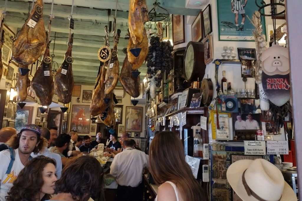 손님이 카운터로 몰려드는 바쁜 타파스 바, 액자 그림으로 장식된 벽, 천장에 걸린 경화 햄.