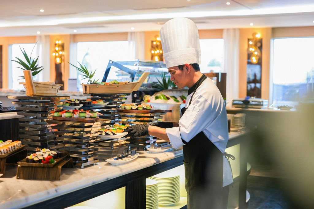 키가 큰 흰색 모자를 쓴 셰프가 조명이 밝은 식사 공간의 뷔페 테이블에 음식을 정리합니다. 접시와 식물도 보입니다.