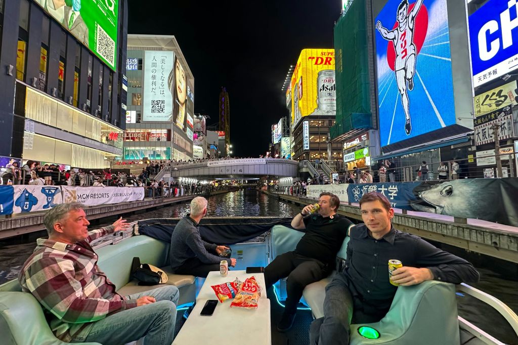 밤에 불이 켜진 광고판으로 둘러싸인 운하에서 보트를 타고 있는 세 사람. 글리코 런닝맨 간판이 특징입니다.