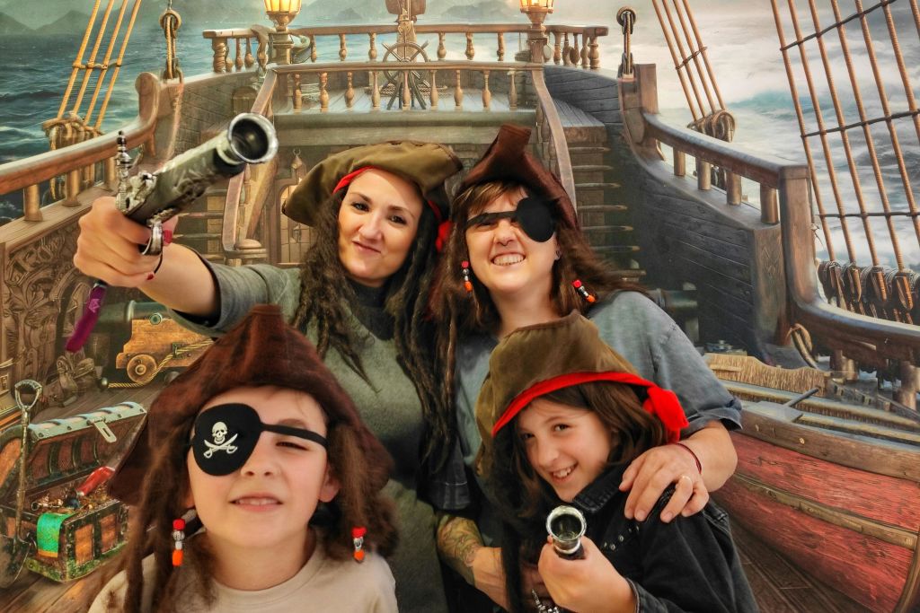 해적으로 옷을 입은 네 명이 모자, 접안경을 쓰고 장난감 무기를 들고 배를 배경으로 포즈를 취하고 있습니다.