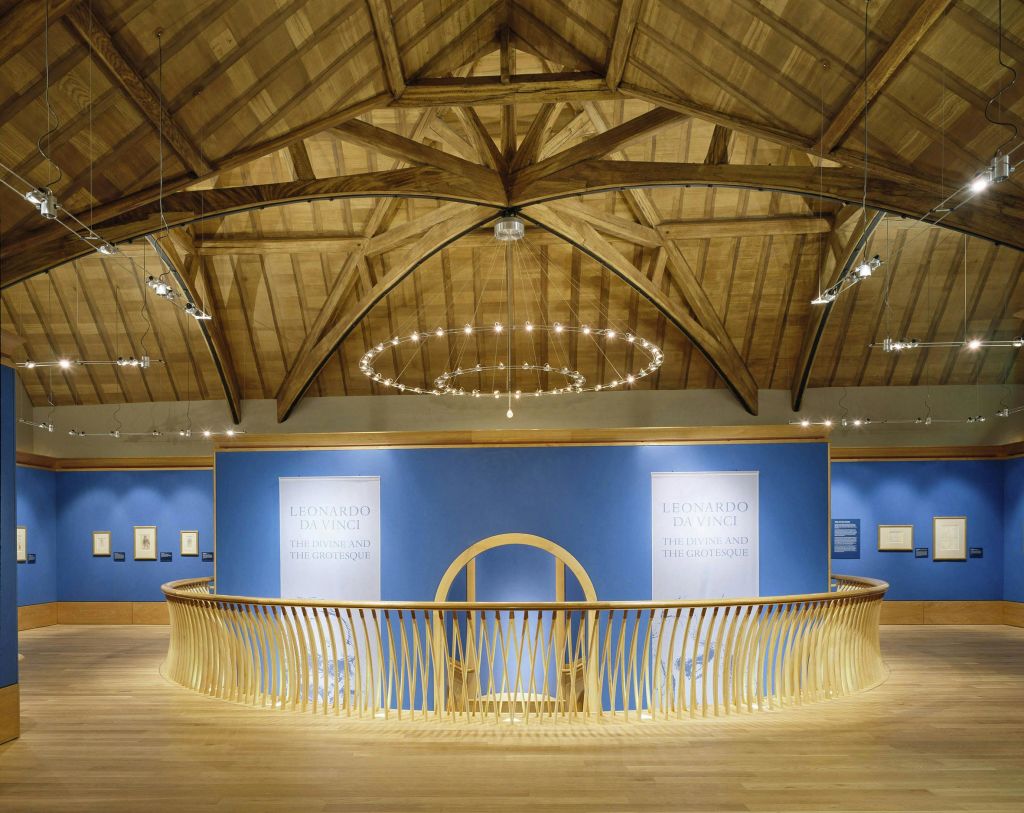 아치형 나무 천장 아래 파란색 벽과 나무 난간, 샹들리에가 있는 박물관 전시관입니다. 표지판에는 '레오나르도 다빈치' 라고 쓰여 있습니다.