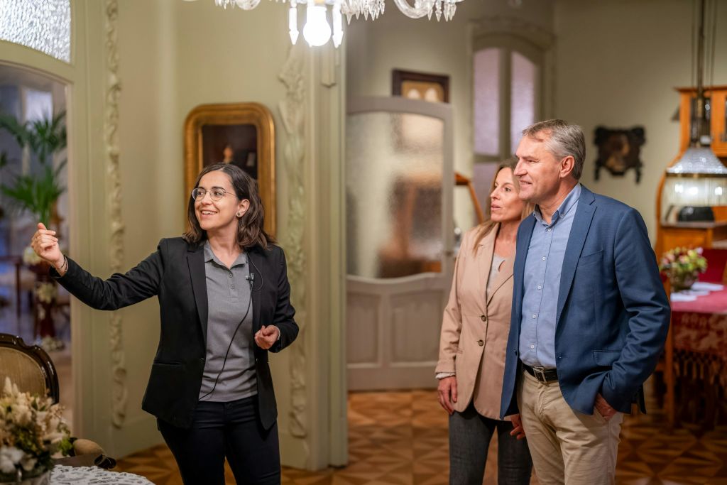 블랙 블레이저를 입은 여성이 샹들리에와 화려한 가구를 포함한 우아한 인테리어가 돋보이는 방에 서서 제스처를 취하고 있습니다.