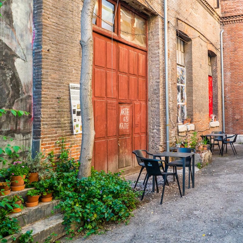 붉은 나무 문, 화분, 나무, 벽돌 벽을 배경으로 한 야외 테이블과 의자가 있는 골목. "Arte Es Arte" 가 문에 쓰여 있습니다.