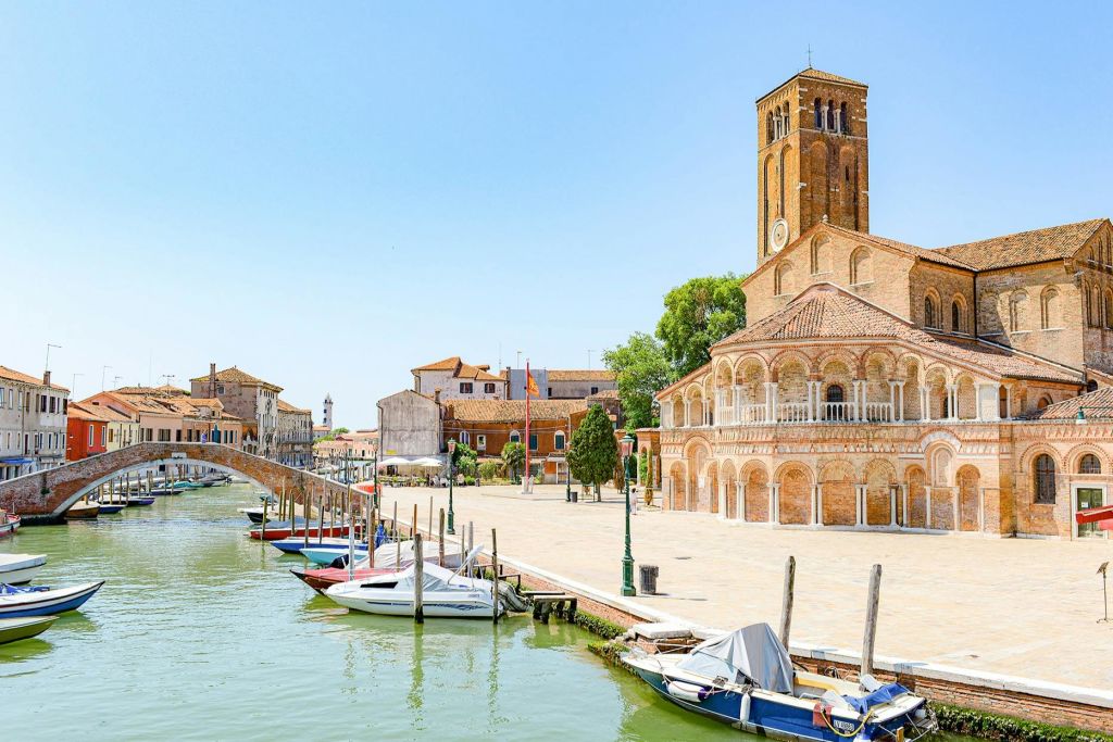 유럽풍 마을의 햇살이 내리쬐는 광장에 보트, 돌다리, 화려한 벽돌 교회가 있는 운하.