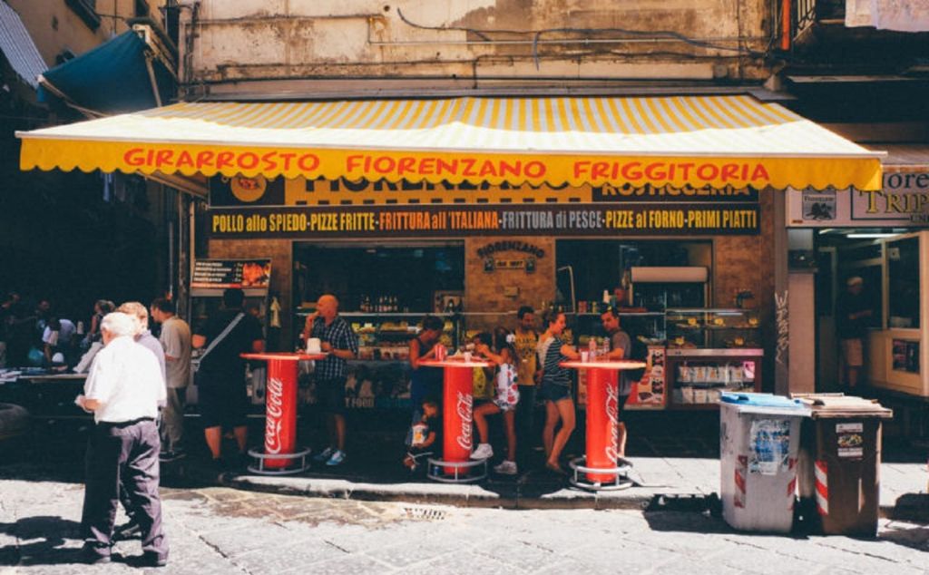노란 줄무늬 천막과 이탈리아 간판이 있는 음식 가판대 바깥의 빨간색 코카콜라 테이블 주변에 사람들이 모여듭니다.