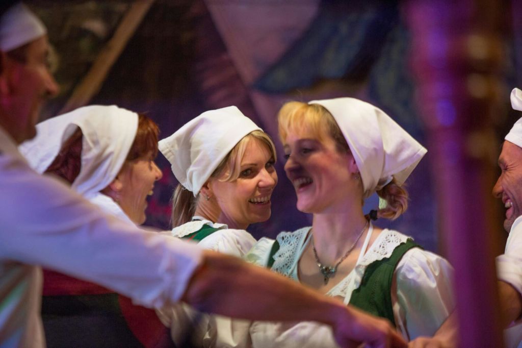 하얀 머리에 스카프를 두르고 전통 의상을 입은 세 명의 여성이 함께 춤을 추며 즐겁게 웃고 있습니다.