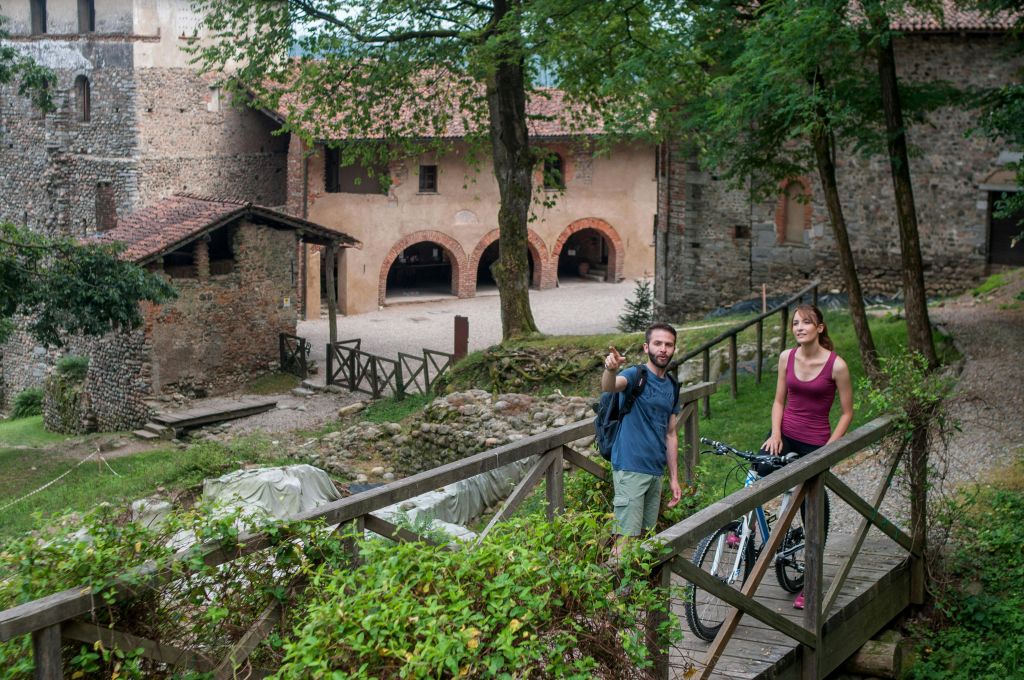 자전거를 탄 두 사람이 유서 깊은 석조 건물과 울창한 녹지 옆의 나무 다리에 서 있습니다.