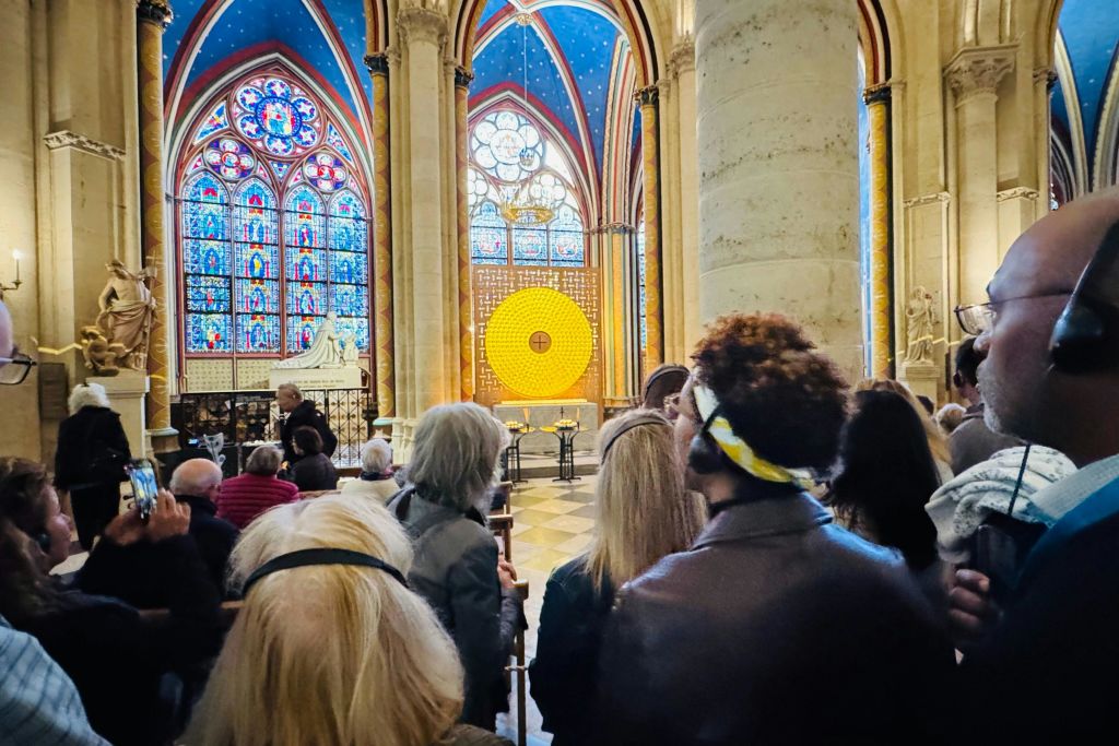 정교한 스테인드글라스 창문과 커다란 원형 노란색 조명이 있는 대성당 내부에 사람들이 서 있습니다.
