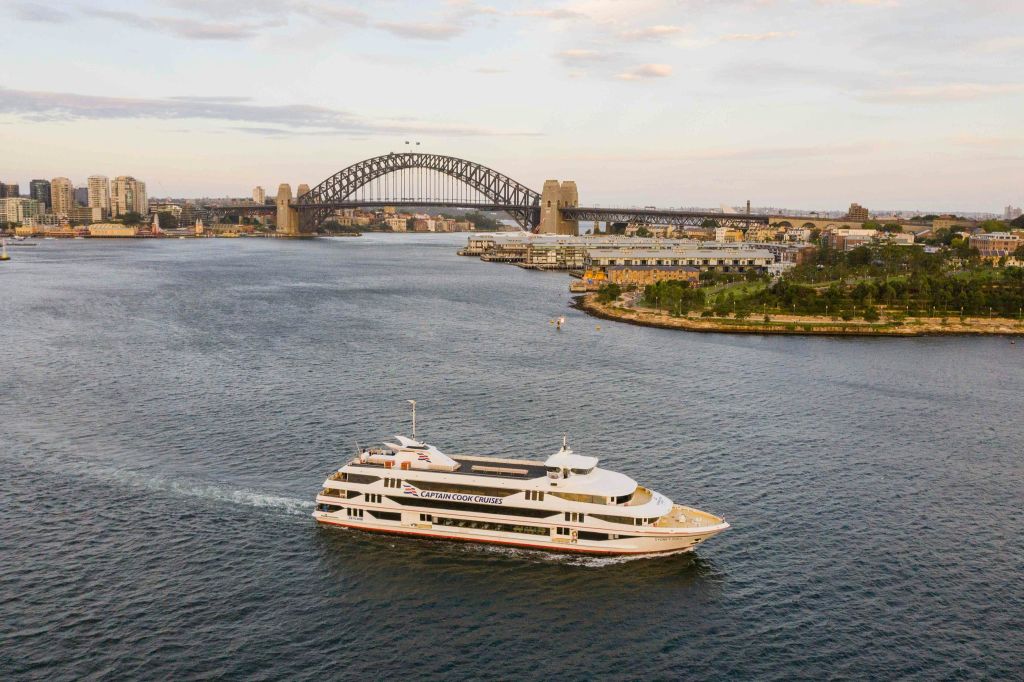 커다란 아치형 다리와 도시 풍경을 배경으로 항구를 항해하는 '캡틴 쿡 크루즈' 라는 이름의 요트.