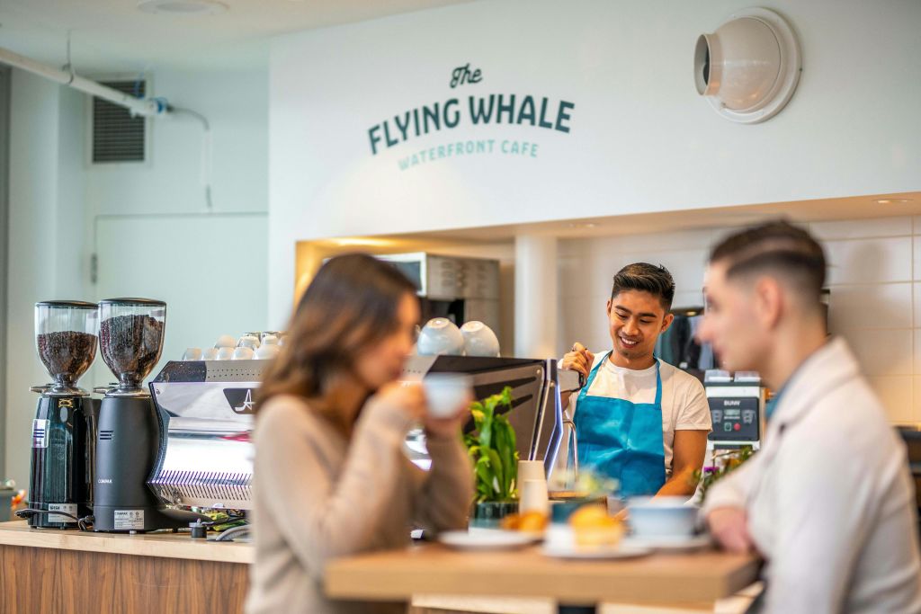 파란색 앞치마를 두른 바리스타가 플라잉 웨일 카페 (The Flying Whale Café) 에서 음료를 제공합니다. 앞에 두 명의 고객이 앉아 있습니다.