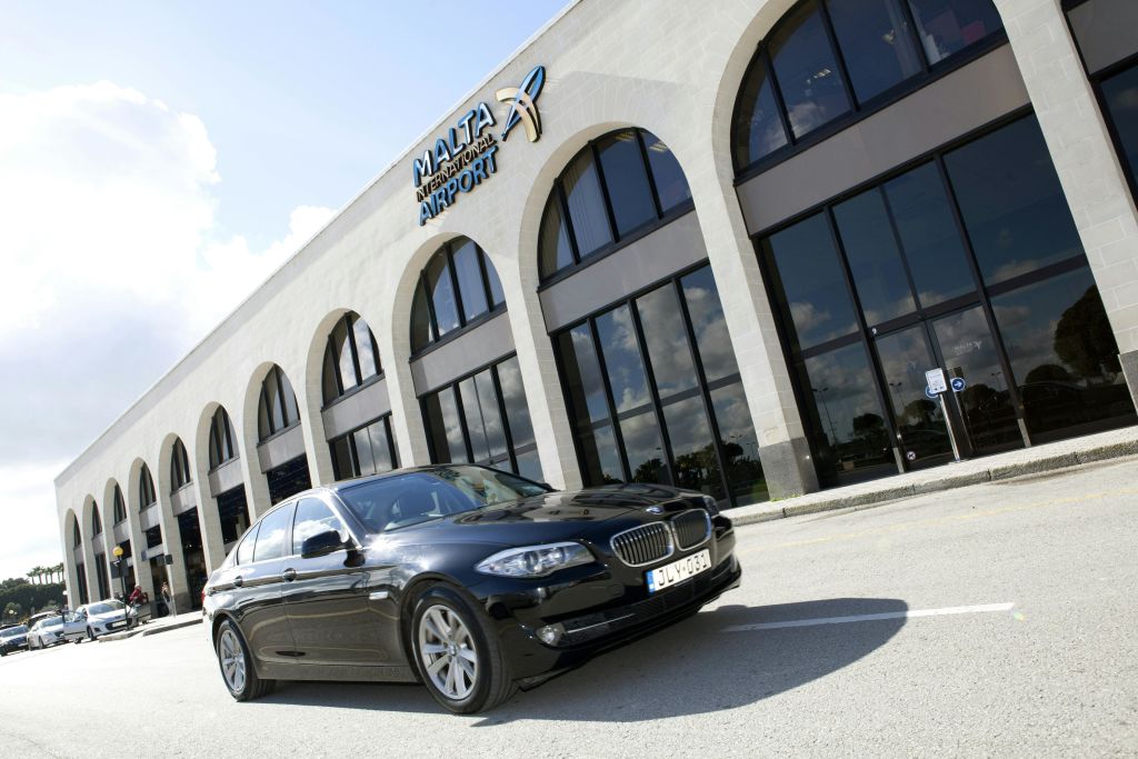 검은색 차량이 몰타 국제공항 건물 앞에 주차되어 있으며, 커다란 아치형 창문과 반사유리가 특징입니다.