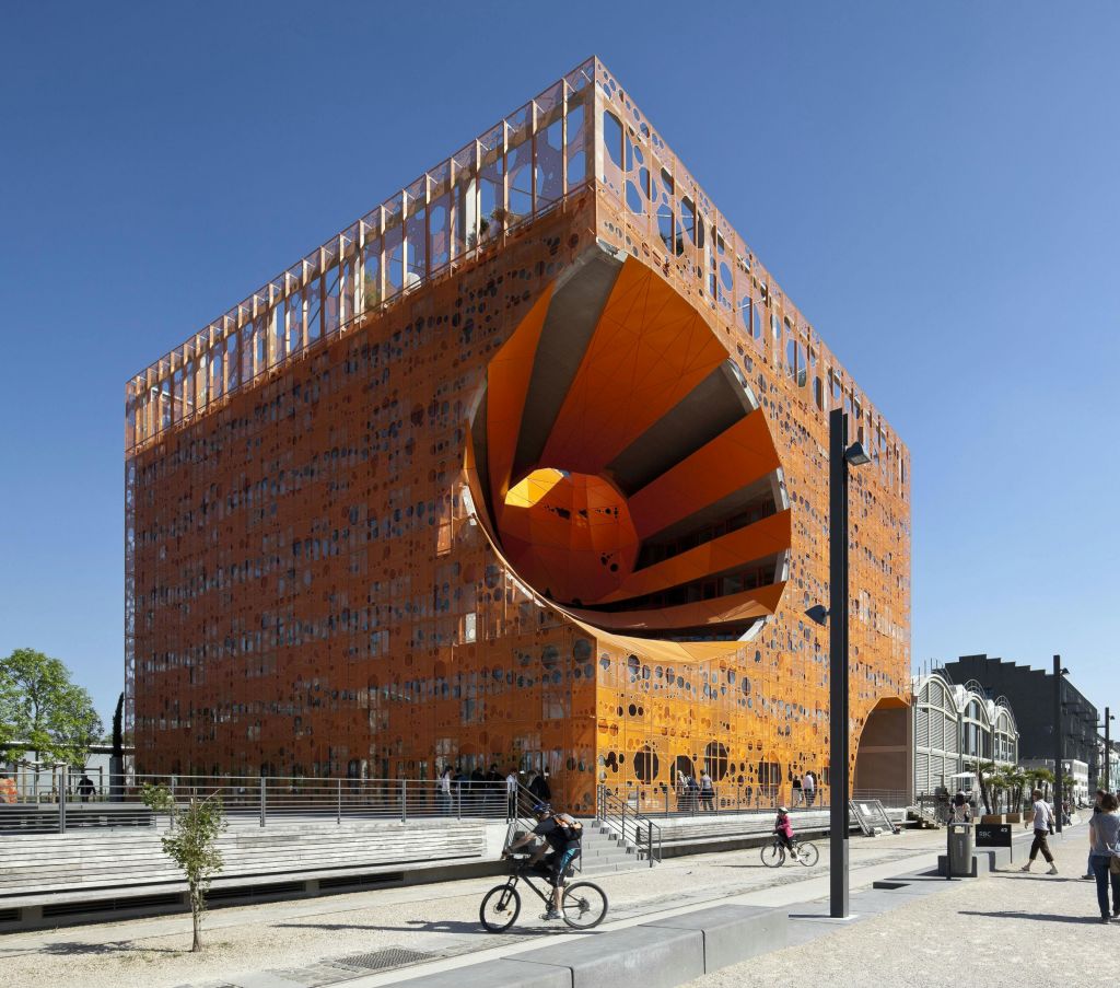 한쪽에 커다란 원형 컷아웃이 있는 현대적인 주황색 큐빅 건물. 사람들은 맑고 푸른 하늘 아래 전경에서 걷고 자전거를 탄다.