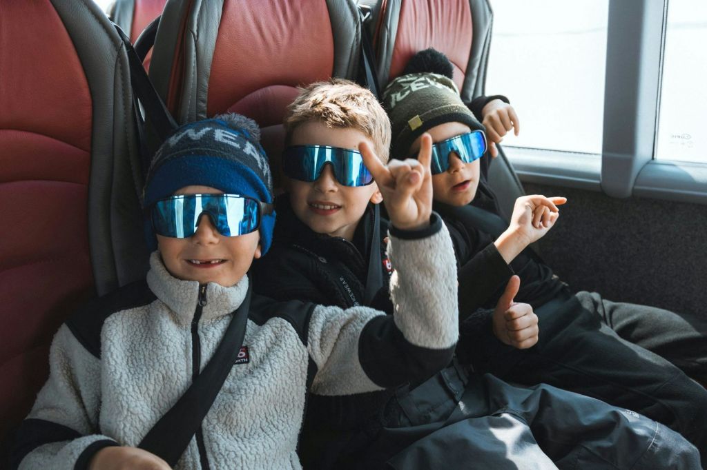 선글라스와 겨울 모자를 쓰고 버스에 앉아 미소를 짓고 손짓을 하는 세 명의 어린이.
