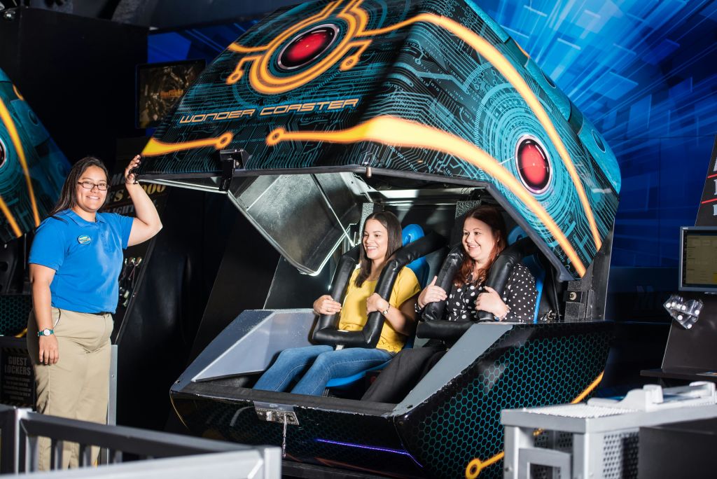 "Wonder Coaster" 브랜딩이 있는 미래형 라이드 시뮬레이터 안에 앉아 있는 두 명의 여성이 제복을 입은 시종 옆에 서 있습니다.