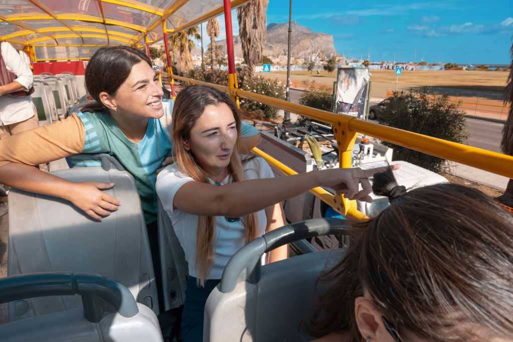 오픈탑 버스의 상부 갑판에 앉아 있는 두 여성이 한 여성은 신나게 가리키고 다른 여성은 미소를 짓고 있습니다.