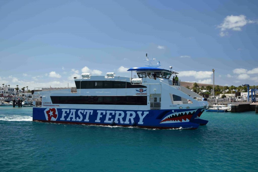 부분적으로 흐린 하늘 아래 맑고 푸른 물에 상어 그래픽 돛이 달린 "빠른 페리" 라는 파란색과 흰색 페리.