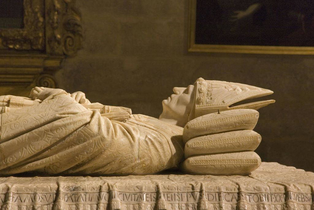 중세 시대의 모습을 형상화한 대리석 무늬가 조각된 석판 위에 놓여 있으며, 세밀한 로브와 주석으로 장식된 교차된 손이 휴식을 취하고 있습니다.