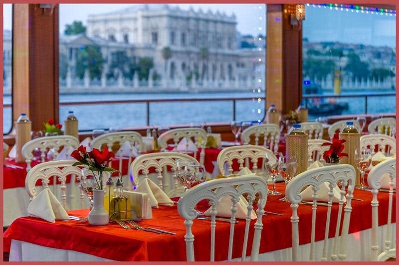 유서 깊은 해안가 건물이 내려다보이는 창문 근처에 빨간색 식탁보, 흰색 의자, 깔끔하게 세팅된 테이블이 있는 우아한 레스토랑.