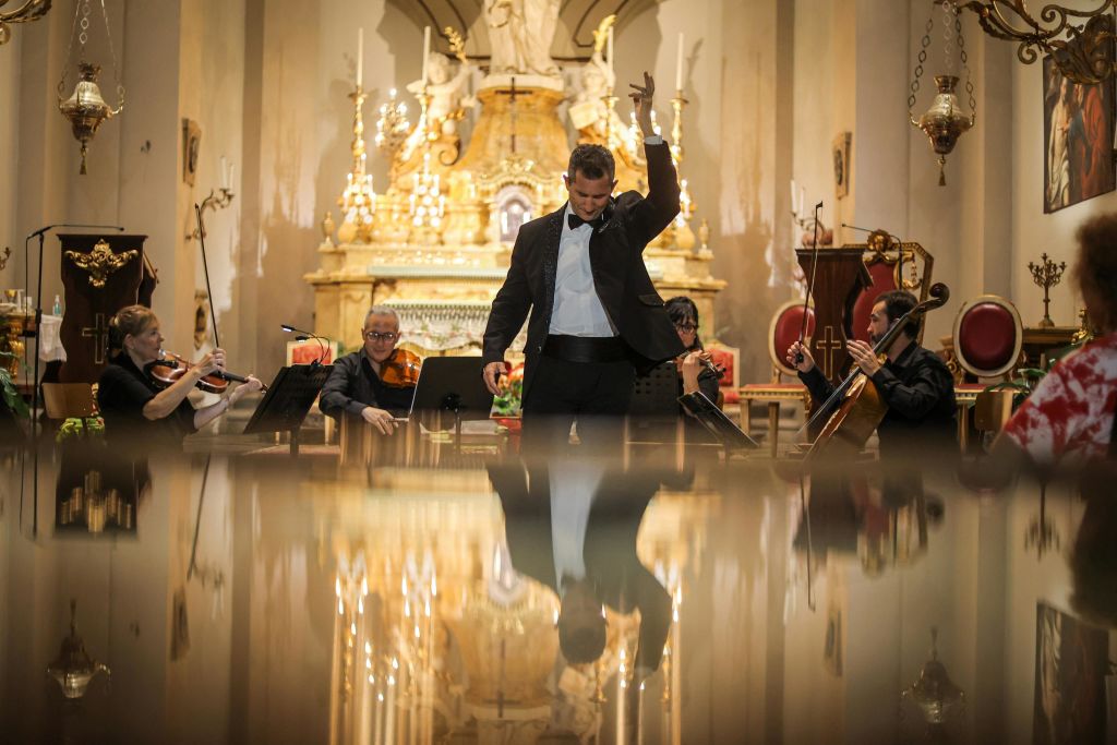 수트를 입은 한 남성이 웅장하고 화려한 교회 내부의 오케스트라 앞에서 극적으로 제스처를 취하고 있다.