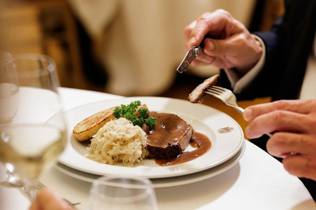 흰색 접시에 으깬 감자, 사우어크라우트, 장식품을 담은 스테이크를 자른 사람이 식탁에 앉았습니다.