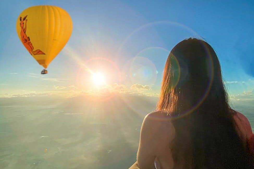 긴 머리카락을 가진 사람이 하늘에서 노란색 열기구가 보이는 높은 전망대에서 일출을 감상합니다.