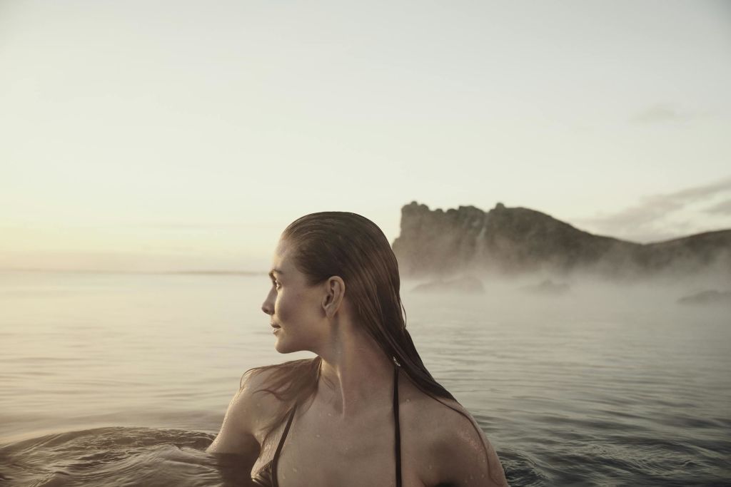 젖은 머리카락을 가진 사람이 물에 부분적으로 잠겨 부드러운 일몰 하늘 아래 바위 해안선을 바라보고 있습니다.