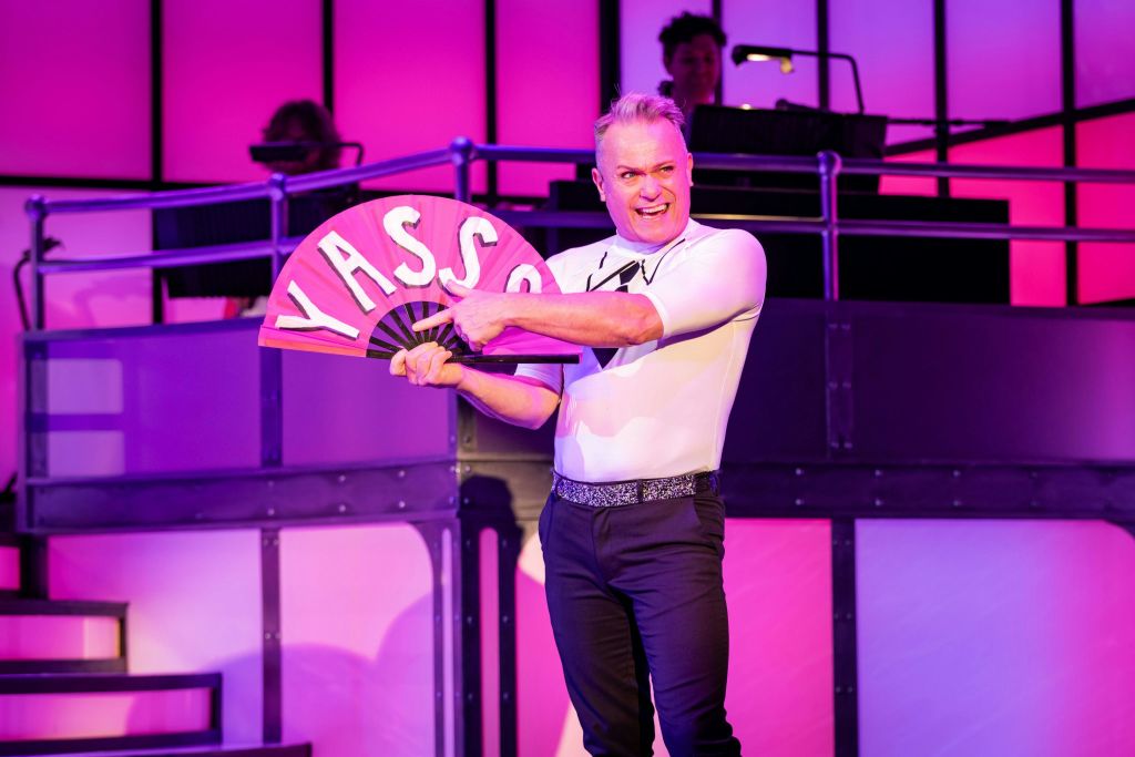 한 남성이 보라색과 분홍색 배경으로 무대에서 공연하는 동안 "YASSS" 라고 적힌 분홍색 팬을 가리키고 있다.