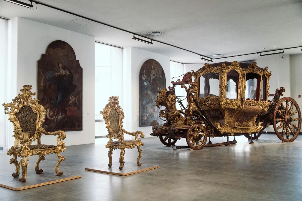 화려한 금색 마차와 두 개의 정교하게 조각된 의자가 전시되어 있으며, 벽 뒤에 커다란 액자 그림이 있습니다.