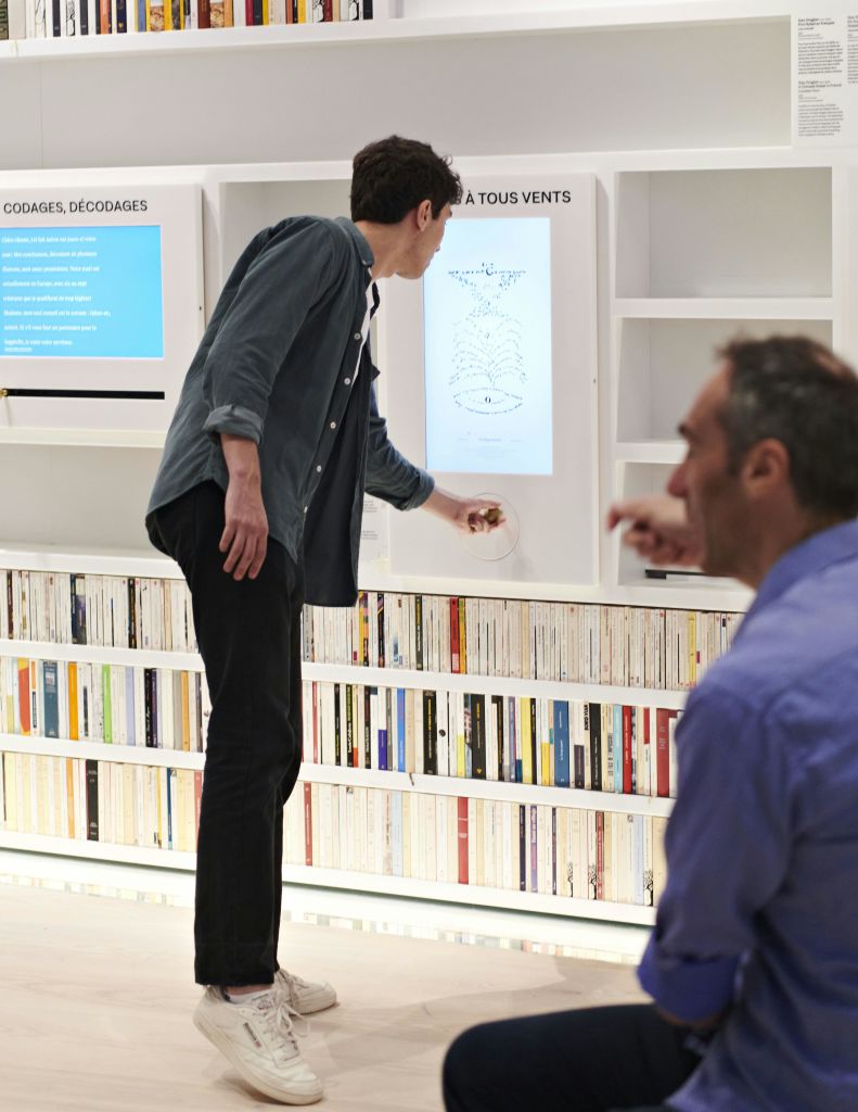 한 남자가 현대 도서관에서 디스플레이와 상호 작용하고 다른 사람은 전경에 앉아 있습니다. 책은 선반에 깔끔하게 정리되어 있습니다.