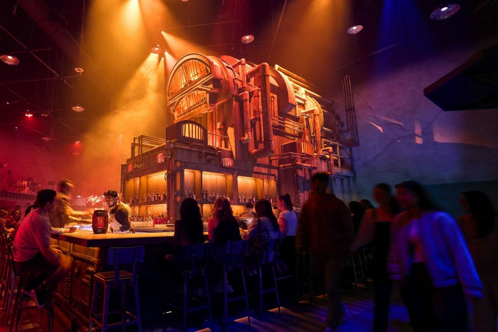 인더스트리얼 테마의 인테리어와 따뜻하고 다채로운 조명이 있는 바에 여러 사람이 앉아 있습니다.