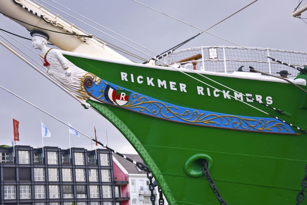 파란색과 금색으로 장식된 "Rickmer Rickmers" 라는 이름의 초록색 배가 회색 하늘 아래 건물 근처에 정박해 있습니다.